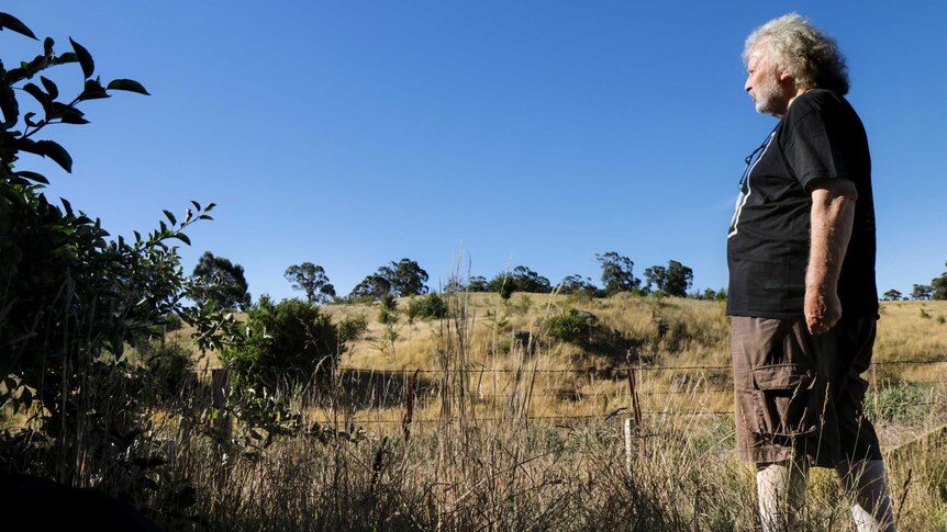 Local John Nightingale wears a black t-shirt, looking out over dry bushland with a blue sky behind.
