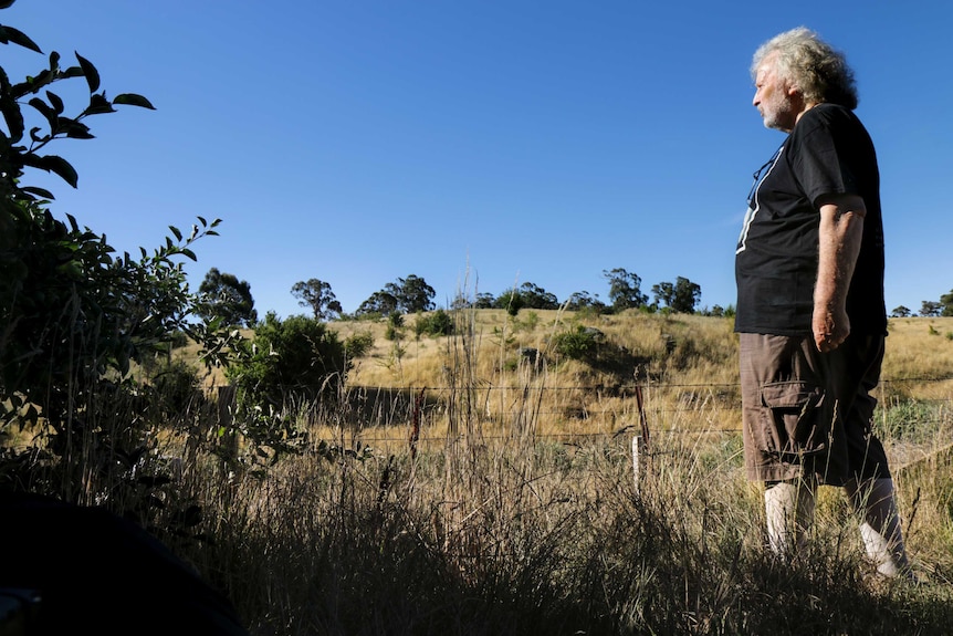 Local John Nightingale wears a black t-shirt, looking out over dry bushland with a blue sky behind.