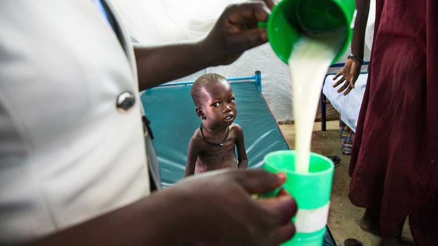 A malnourished child with visible ribs and a distended belly watches an aid worker pour milk. Juba, South Sudan.