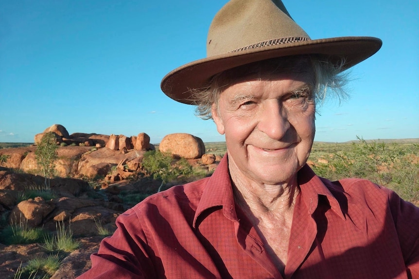 A man in his 70s with a broad hat stands in front of some boulders in the outback, with blue sky overhead.