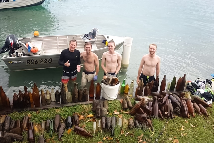 Four men in swimming gear smile after clearing dozens of glass bottles from a clear blue river.