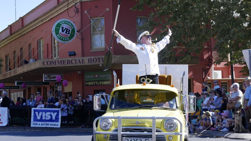 A man stands on a car float with his arms in the air. He wears a white jacket.