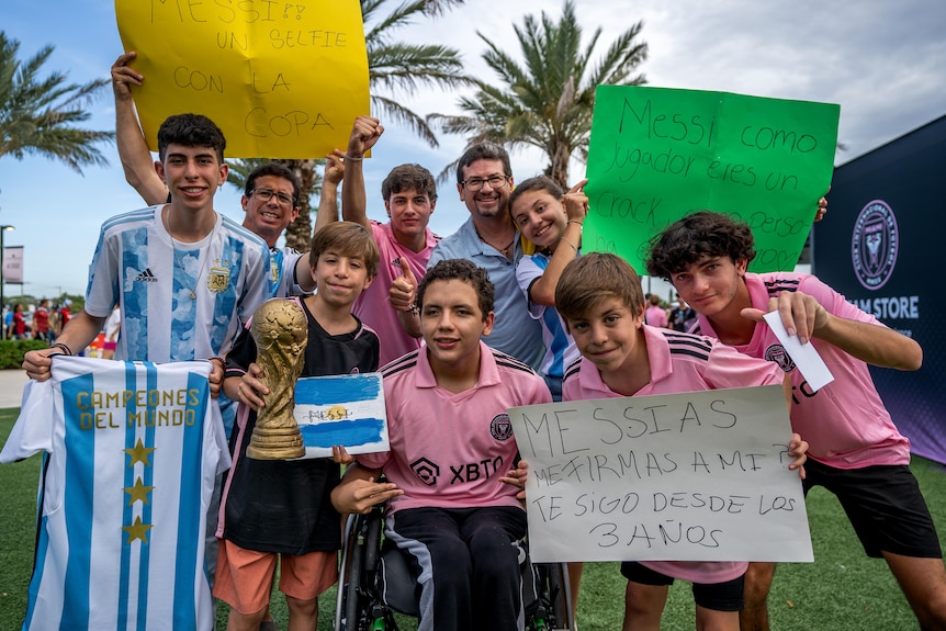 A group of young children with jerseys and signs with messages for Lionel Messi smile together
