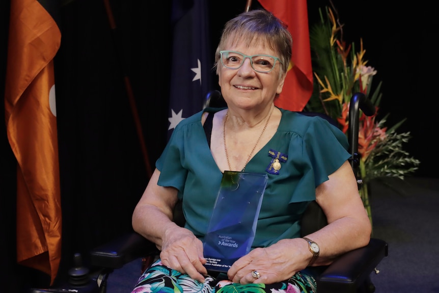 An elderly woman in blue-framed glasses and a blue dress sits in front of the Australian flag and, holding an award.