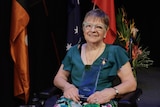 An elderly woman in blue-framed glasses and a blue dress sits in front of the Australian flag and, holding an award.