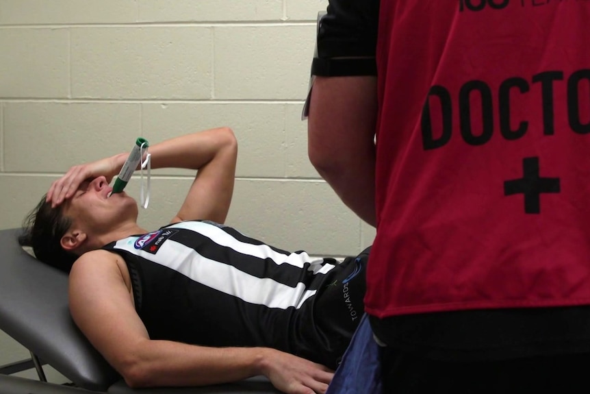 An AFLW player sucks pain relief on a bed after a knee injury.