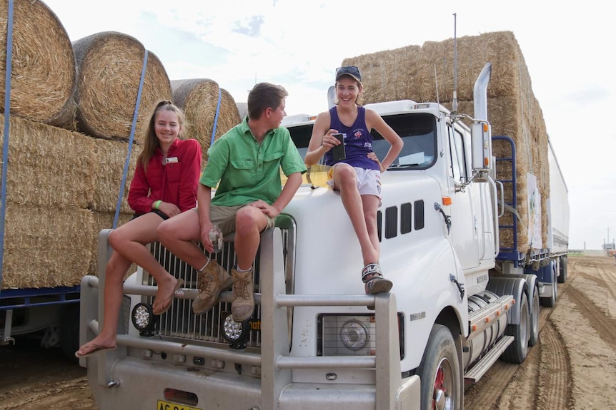 The teenagers sit laughing on the front of a truck loaded with hay.