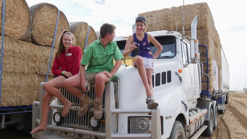The teenagers sit laughing on the front of a truck loaded with hay.