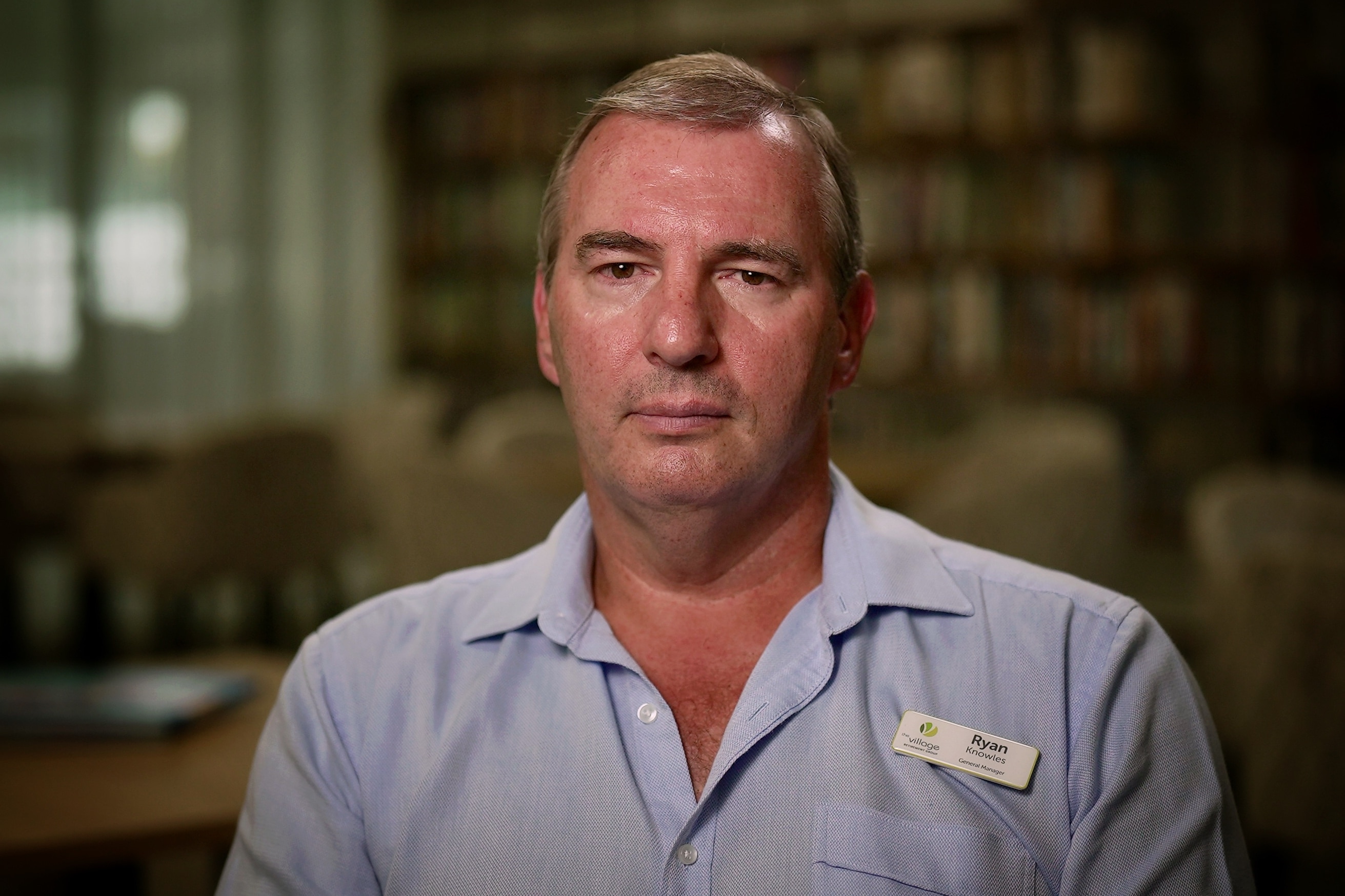 Un hombre blanco, rubio, pelo corto, vestido con una camisa azul con botones y estanterías detrás de él en la biblioteca.