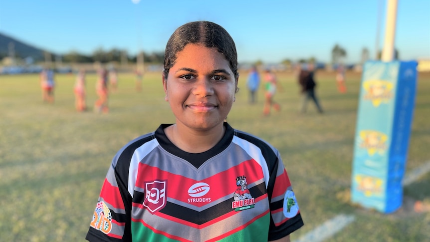 A 14-year-old girl wears a football uniform and stands in front of her local footy field before her NRLW game. She's smiling. 