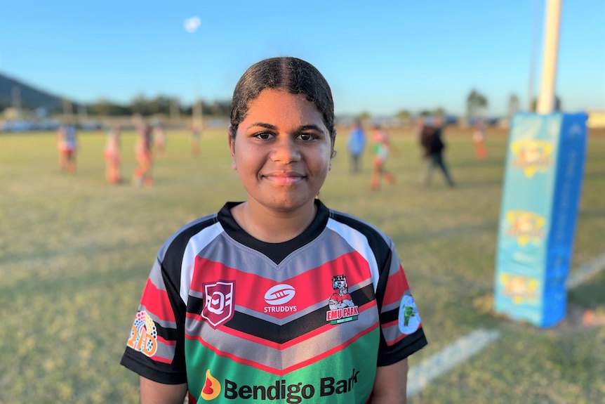 A 14-year-old girl wears a football uniform and stands in front of her local footy field before her NRLW game. She's smiling. 