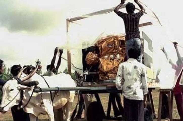 An old photo showing a gold metallic satellite on a cart being pulled by a bullock. 