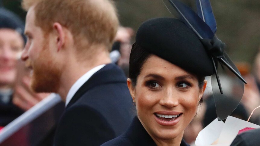 Meghan Markle wears a navy blue fascinator and looks to the camera with a smile on her face with Prince Harry behind her.