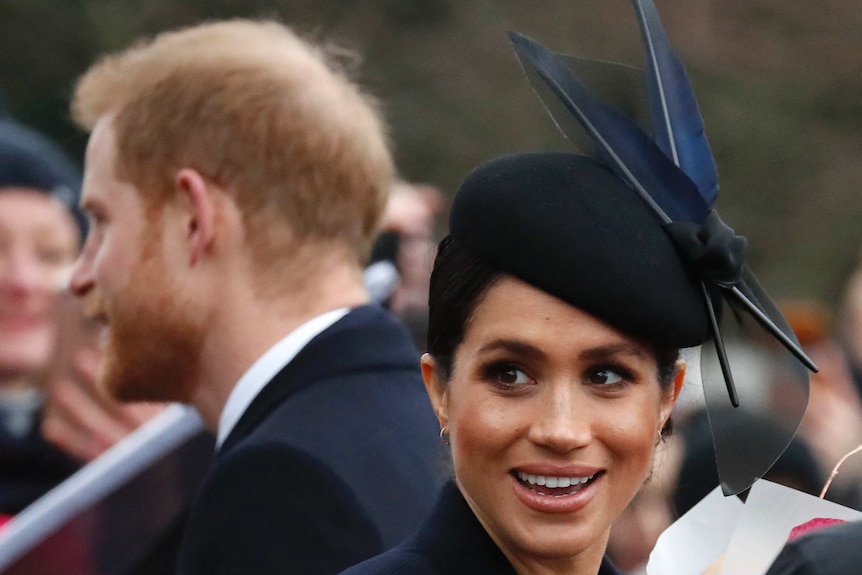 Meghan Markle wears a navy blue fascinator and looks to the camera with a smile on her face with Prince Harry behind her.