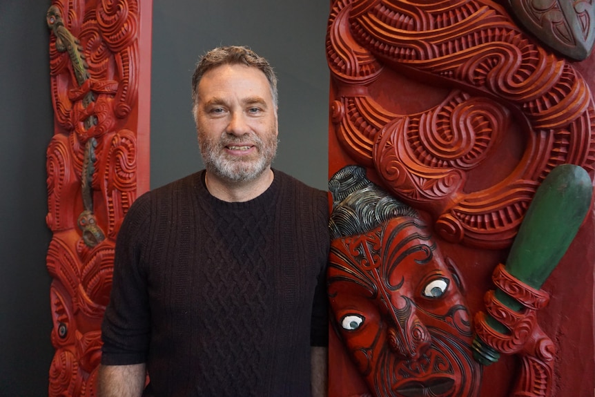 Carwyn is standing in front of a bright red pou with ancestral carvings. He is wearing a black jumper 