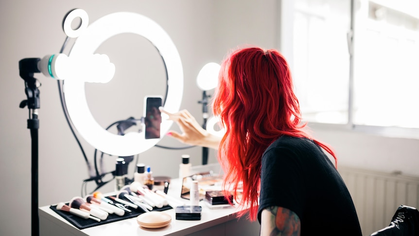 A woman with red hair sits in front of a ring light.