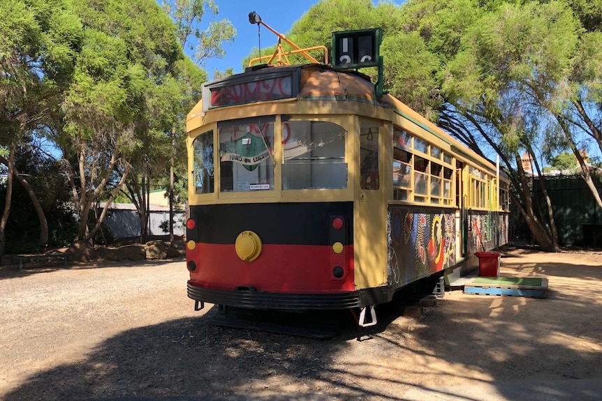 A repurposed tram painted with murals sits in a gravel yard surrounded by trees.