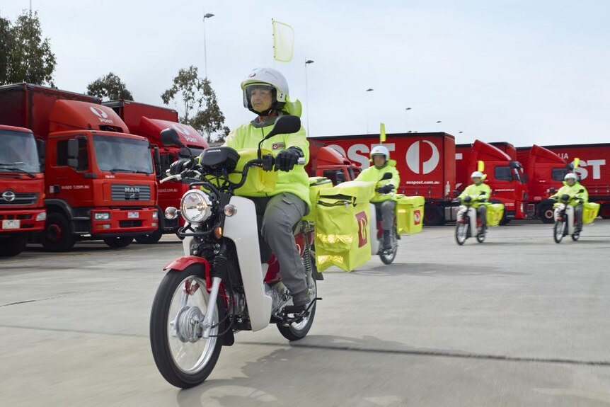 Five Australia Post posties ride out of a carpark on their delivery bikes, with red mail trucks in the background.