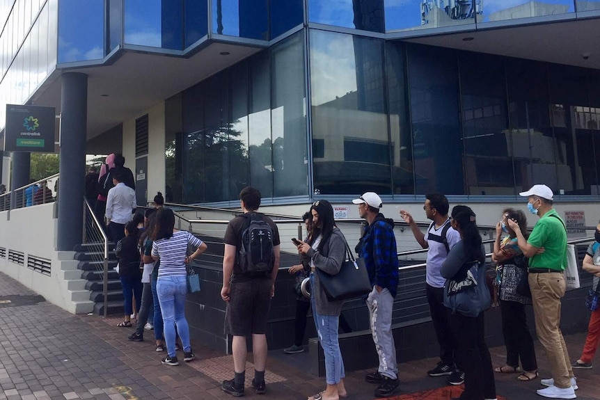 People stand in a line outside a building covered in blue glass