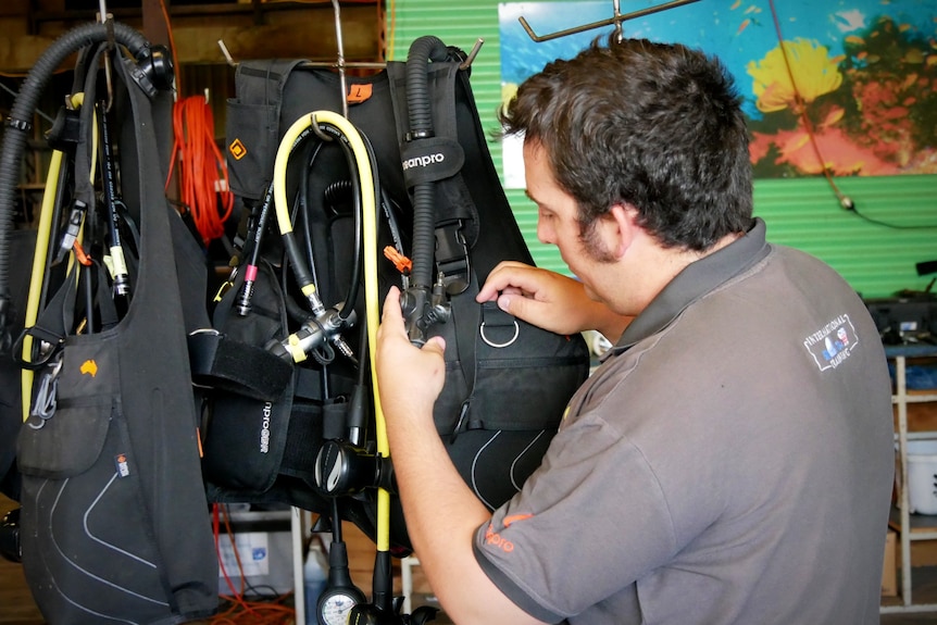 A dark haired man inspects a diving vest, a green ocean background. 