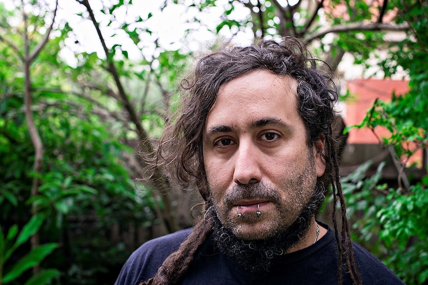 Portrait of a man with two lip piercings, dark brown eyes, and brown and grey dreads.