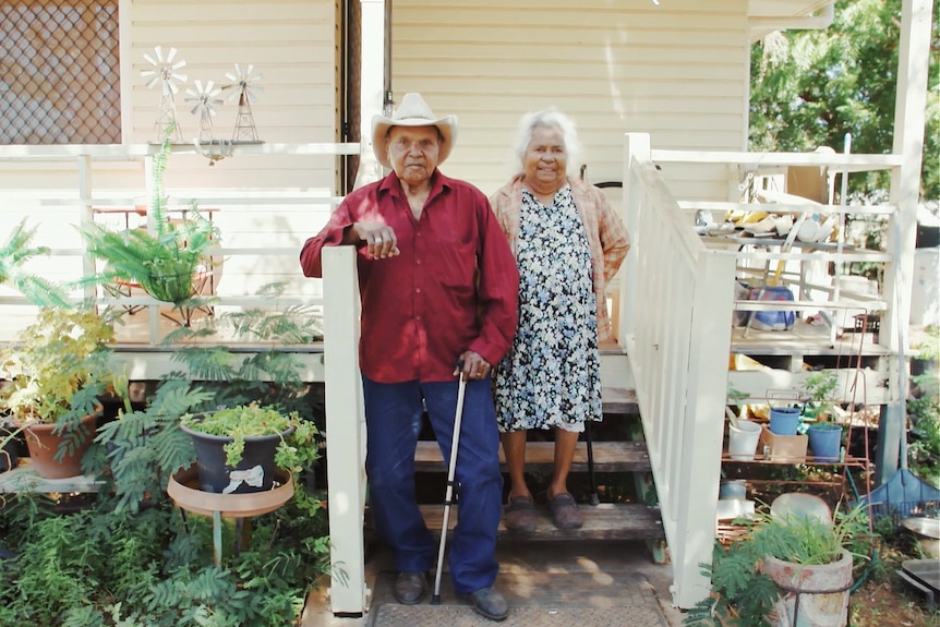An elderly Indigenous couple stand on their front porch of a small wooden house