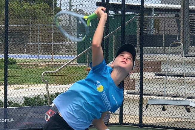 A teenage girl wearing blue t-shirt, shorts, cap, hits a serve on the tennis court.