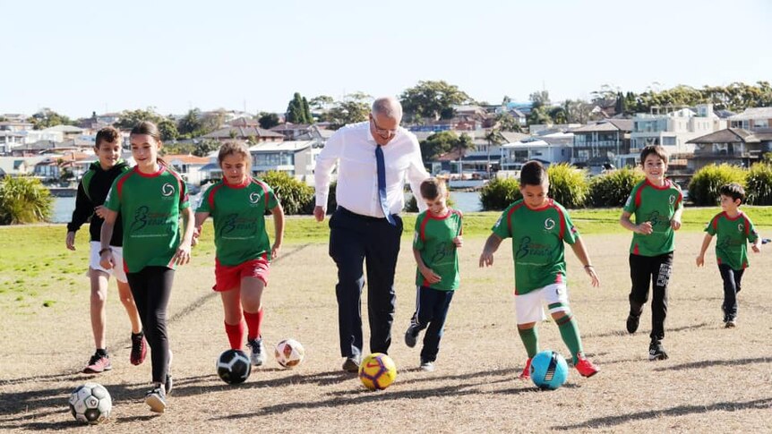 Scott Morrison dribbles a soccer ball alongside children on a suburban soccer field