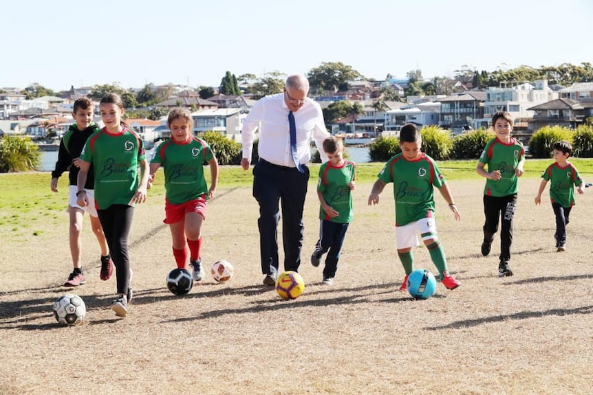 Scott Morrison dribbles a soccer ball alongside children on a suburban soccer field