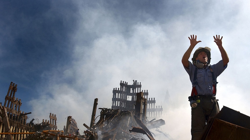 You look up at a fireman as he raises his hands with open palms as stands in front of rubble and a cloud of smoke.