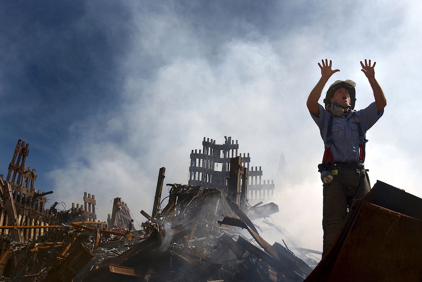 You look up at a fireman as he raises his hands with open palms as stands in front of rubble and a cloud of smoke.