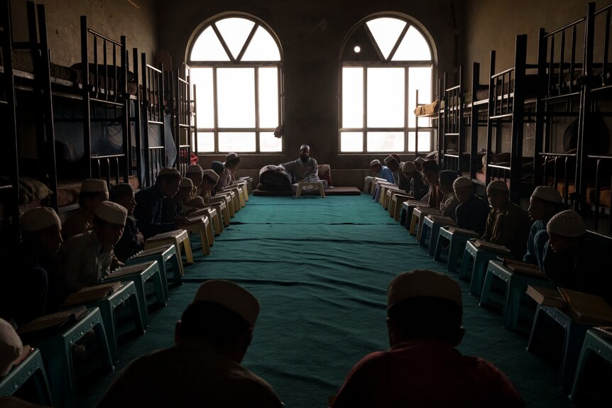 Boys sit in parallel rows facing each other with a teacher in the middle, reading the Quran.