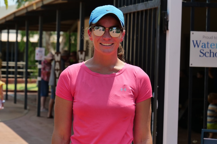 A woman in a pink shirt and blue hat stands outside