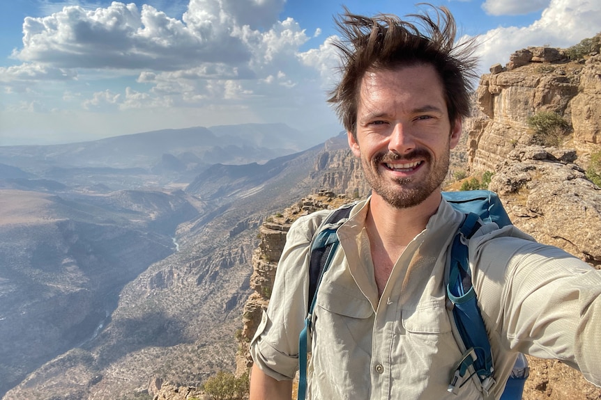 A middle-aged man with brown hair and stubble takes a selfie on top of a high mountain range in Iraq. 