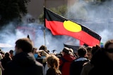 An Aboriginal flag flies at a Perth rally with smoke in the background.
