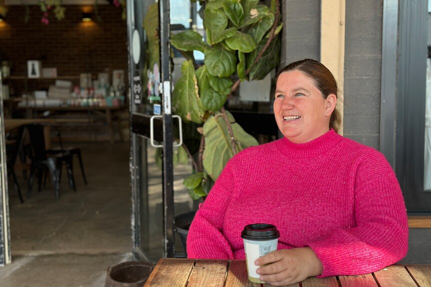 Una mujer sentada afuera de una cafetería sonriendo con una taza de café para llevar en la mano