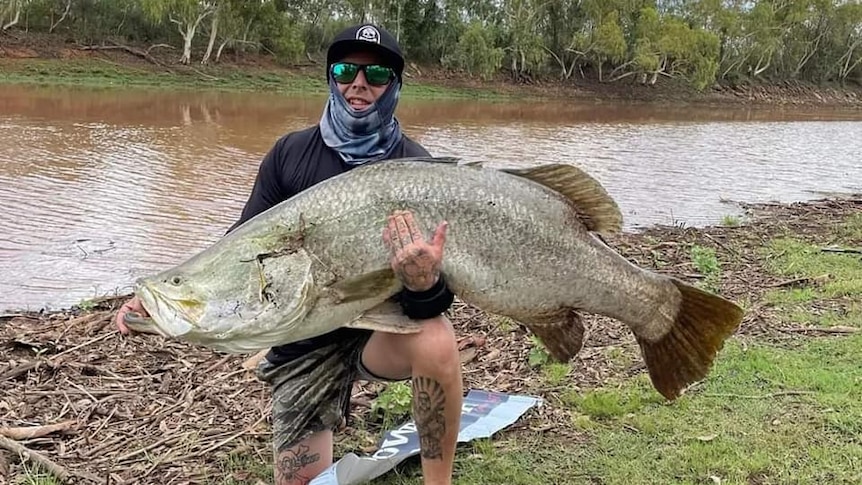 Man in shirt and shorts and navy cap with polarised sunnies on kneels by river holding large barramundi fish