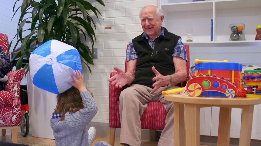 An elderly man sits on a chair playing with a young toddler.