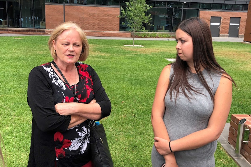 A lady with crossed arms stands next to a younger woman on a green lawn as they speak to the media.