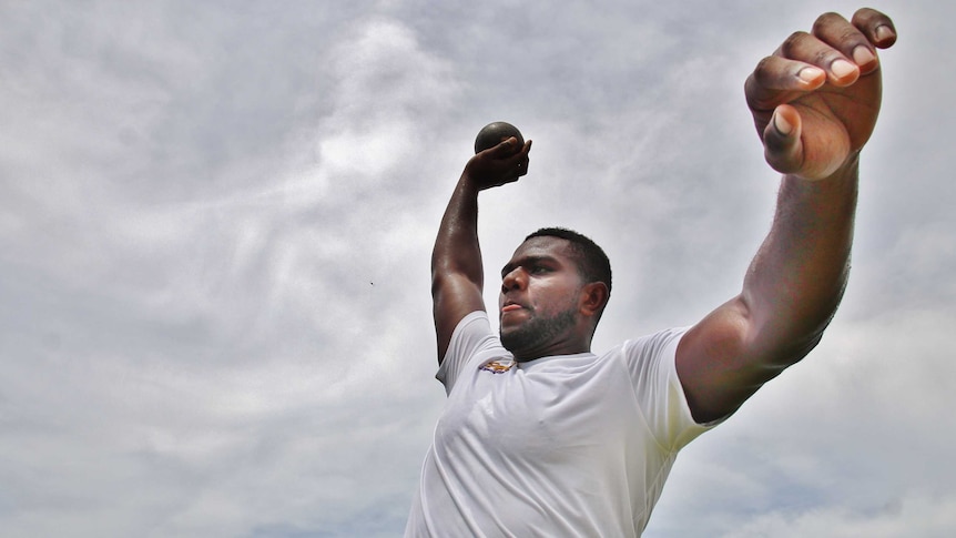 Mustafa holds the shot put in the air with his right hand and raises his left. The sky is behind him.