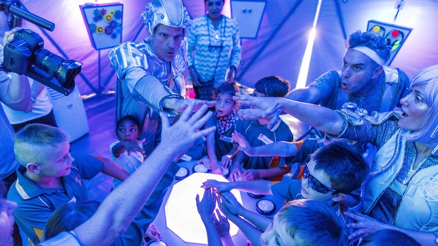 A group of performers in space costumes put their hands in a circle with small children, in a tent of blue and pink light