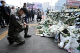 A man in army uniform with a South Korean flag kneeling in front of a pile of flowers