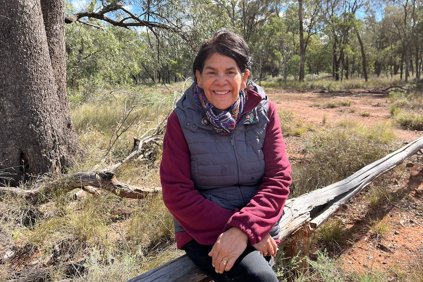  An older woman in a red top, purple puffer jacket, scarf, smiles, sits on a log in a forest, trees, red earth.