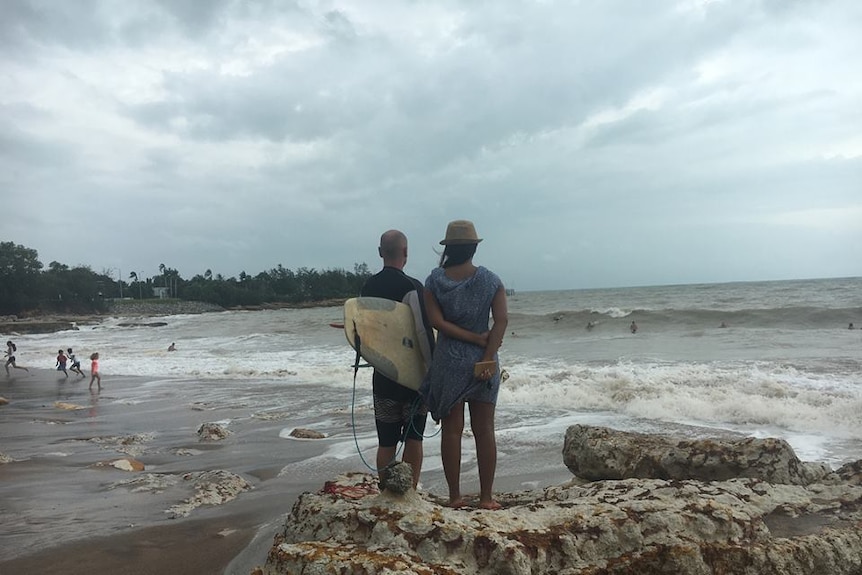 A man holding a surfboard and a woman stand on Nightcliff beach looking at surfers in the waves.