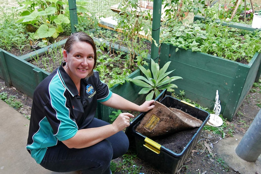 A woman sits beside a worm farm and raised garden beds which have seedlings growing.