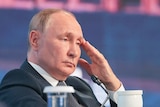 Vladimir Putin sits with a sad expression with his hand on his face infront of a microphone.