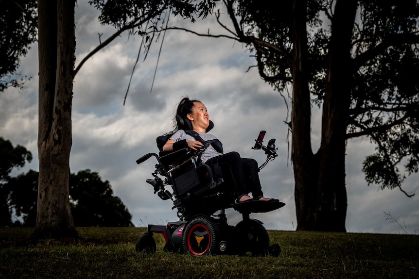Melanie Tran sits in her wheelchair in a park, she looks out into the distance.