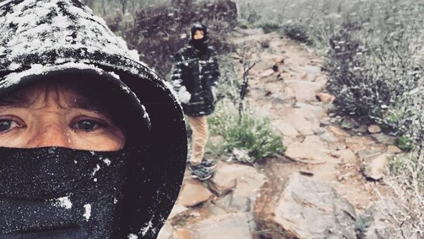 A person with a hood on their head and scarf over their mouth takes a selfie on Bluff Knoll in snow with another person behind.