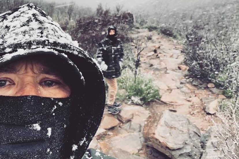 A person with a hood on their head and scarf over their mouth takes a selfie on Bluff Knoll in snow with another person behind.