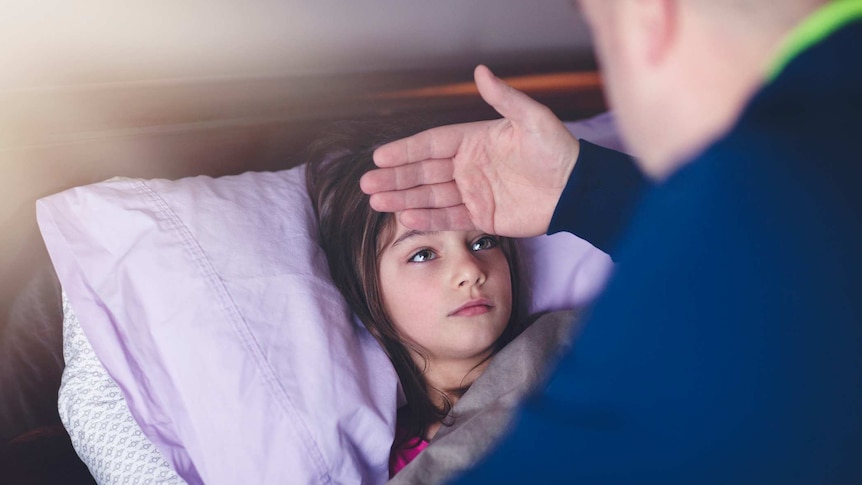 Profile of man in blue jumper leaning over young girl in bed, back of his hand placed on girl's forehead to check fever.